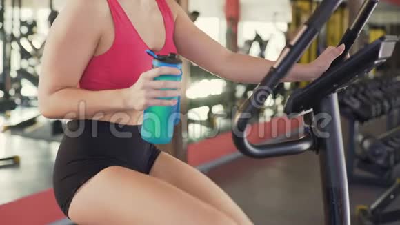 健身房健身苗条的女人一边喝水一边骑健身自行车视频的预览图