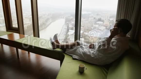 早上入住豪华酒店年轻英俊的年轻商人打电话的后景坐在全景窗附近的沙发上视频的预览图