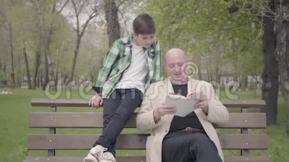 肖像可爱的祖父和可爱的孙子坐在公园的长凳上老人为男孩读这本书视频的预览图