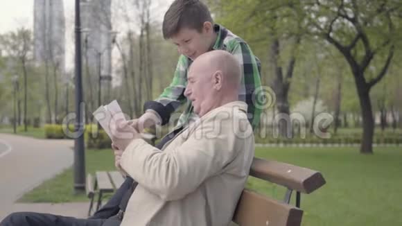 肖像可爱的祖父和可爱的孙子坐在公园的长凳上老人为男孩读这本书视频的预览图