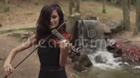 穿着黑色裙子拉着瀑布在森林里拉小提琴的漂亮女孩视频的预览图