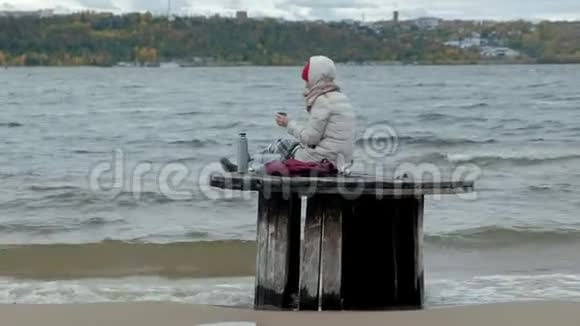 年轻女子穿着暖和的衣服坐在海边坐在木盘管上用热水瓶喝热茶视频的预览图
