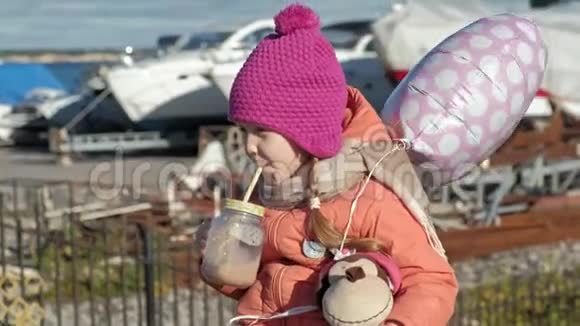 小女孩戴着粉红色的帽子带着气球坐在河港旅行微笑喝可可饮料视频的预览图