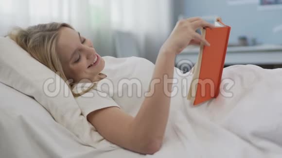 笑着高兴的少女在看一本关于浪漫爱情的小说躺在床上视频的预览图