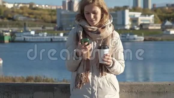 年轻的女人穿着白色夹克在河港喝咖啡用电话微笑特写视频的预览图