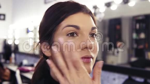 化妆师在一个漂亮的深褐色头发的女孩的脸上涂上粉底的过程视频的预览图