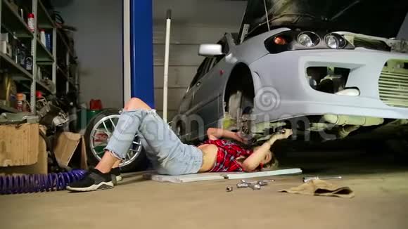 年轻的金发女工穿着格子衬衫和破牛仔裤躺在拆卸的汽车下面修理汽车服务视频的预览图