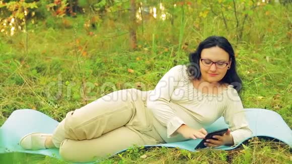 森林里一个戴眼镜的年轻女子躺在地毯上做瑜伽在平板电脑上用信使传递信息视频的预览图