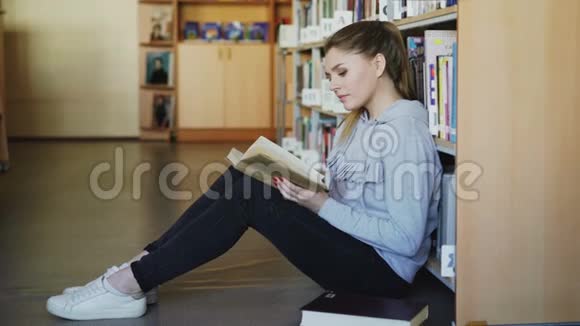 穿着休闲服的年轻漂亮的白人女学生正坐在书架旁宽敞的大图书馆地板上视频的预览图