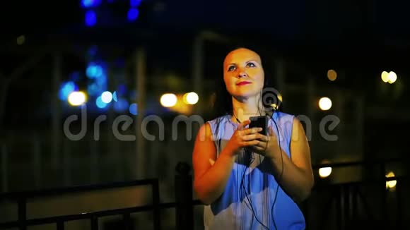 年轻的笑女在码头戴着耳机听着电话里的音乐在夜空中唱歌视频的预览图