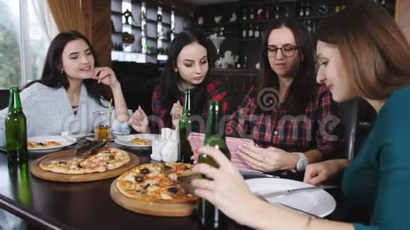 四个快乐的女朋友在一家餐馆吃披萨喝啤酒看平板电脑上的照片视频的预览图