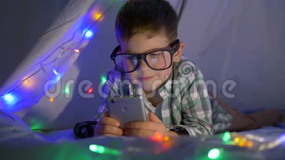 带眼镜的小男孩在帐篷里用手机看视频家里有花环视频的预览图
