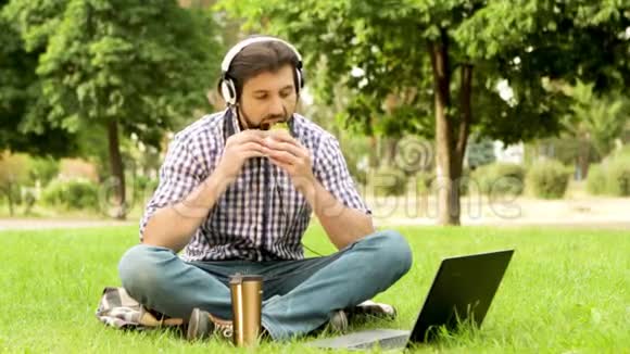 快乐的家伙坐在草地上看着笔记本电脑他在听音乐和微笑他在咬人视频的预览图