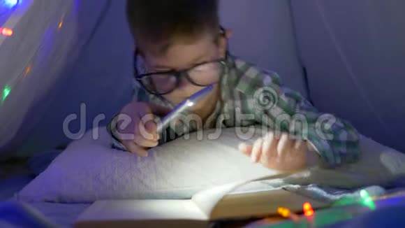 在家上学戴眼镜的男孩拿着一部智能手机手里拿着手电筒在帐篷里看书视频的预览图