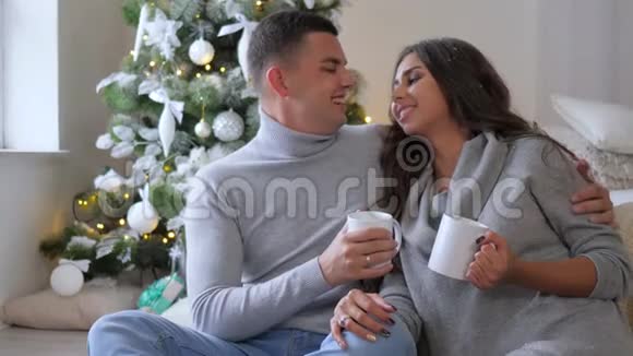早上在圣诞树的背景下丈夫和妻子手里拿着白色的大杯子在家里的沙发上视频的预览图