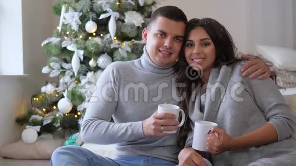 可爱的夫妇带着白色的杯子早上在装饰圣诞树的背景下看沙发上的摄像机视频的预览图