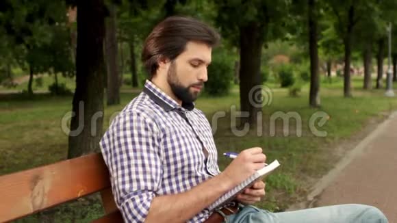 严肃的家伙坐在公园的长凳上在笔记本上写笔记他看起来很严肃男人正在喝一杯咖啡视频的预览图