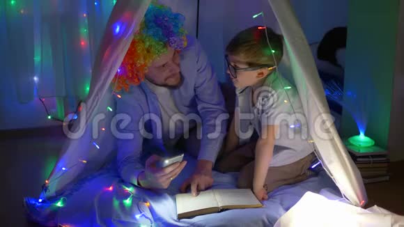 小男孩和小丑一起玩坐在装饰着明亮花环的帐篷里读童话视频的预览图