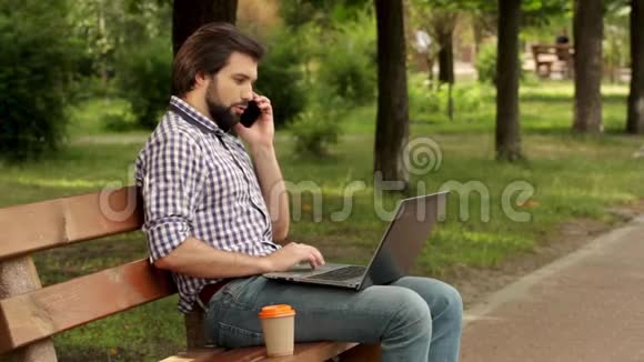 男人坐在长凳上打电话他腿上拿着笔记本电脑人拿着白色手机看着他是视频的预览图