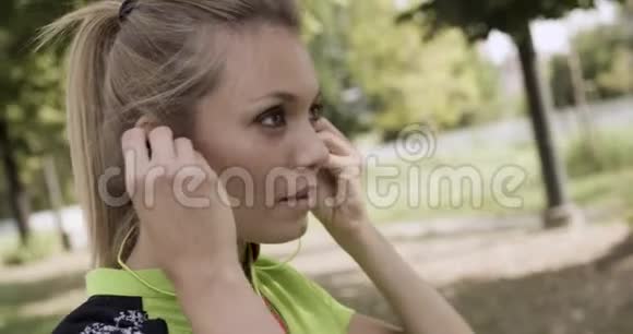 穿着健身服的金发女青年戴着耳机进行跑步训练夏日阳光明媚工业绿城城市视频的预览图