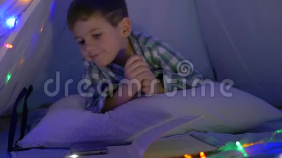 戴眼镜的小男孩看着相机晚上在帐篷里用手拿着手机视频的预览图