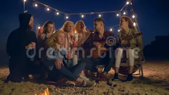 年轻人用吉他唱歌晚上在沙滩上坐在篝火旁喝塑料杯里的热饮视频的预览图