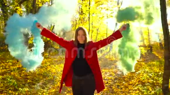 穿着红色外套的漂亮女孩手里拿着彩色的烟走过黄色的秋林视频的预览图