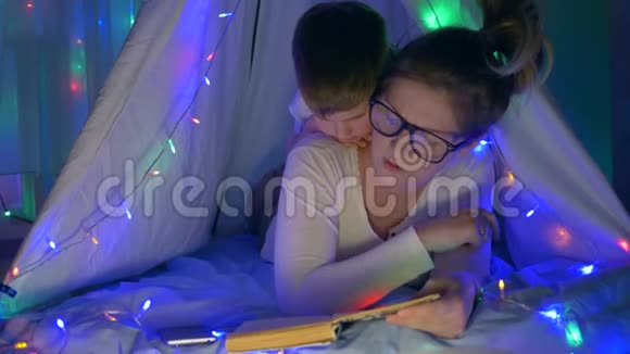母亲的关怀年轻的母亲和儿子在帐篷里看书室内有明亮的节日视频的预览图