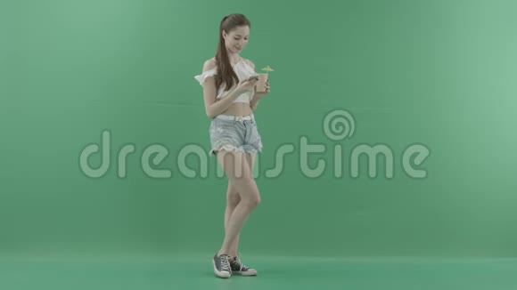 性感的年轻女性在绿色背景下喝鸡尾酒时在智能手机上发短信视频的预览图