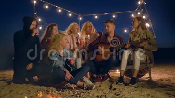 夜晚的海滩一群带着吉他唱歌的年轻人在沙滩上坐在篝火旁喝着塑料杯里的热茶视频的预览图