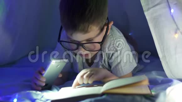 夜间阅读戴眼镜的可爱的孩子在黑暗的室内在手电筒下的杂志上看到视频的预览图