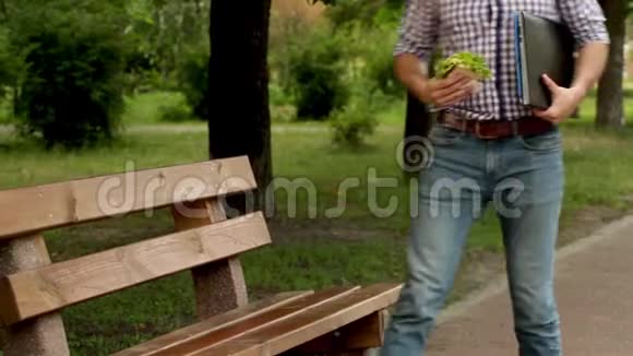 男人走到长凳上坐在上面他把文件夹和汉堡放在长凳上男人把笔记本放在腿上打开它盖盖视频的预览图