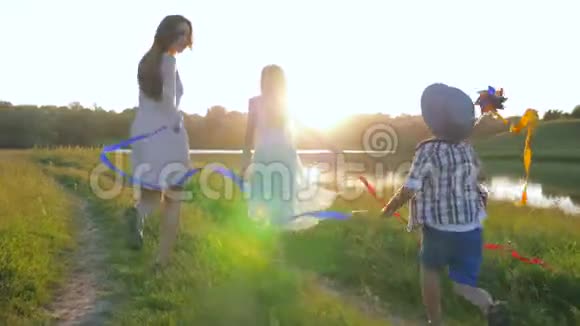 家庭娱乐明亮的孩子们带着彩带在明亮的阳光下沿着河边的绿色草地奔跑视频的预览图