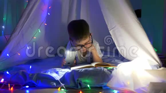 阅读书籍聪明的孩子戴上眼镜在手电筒的灯光下阅读童话故事躺在帐篷里戴着花环视频的预览图