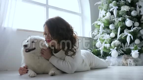 舒适的寒假美丽的女孩抱着狗躺在地板上靠近装饰圣诞树视频的预览图