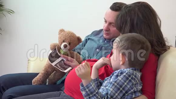年轻的父亲怀孕的母亲小儿子和玩具熊玩具等待新家庭玩具抚摸女人的肚子孩子准备视频的预览图