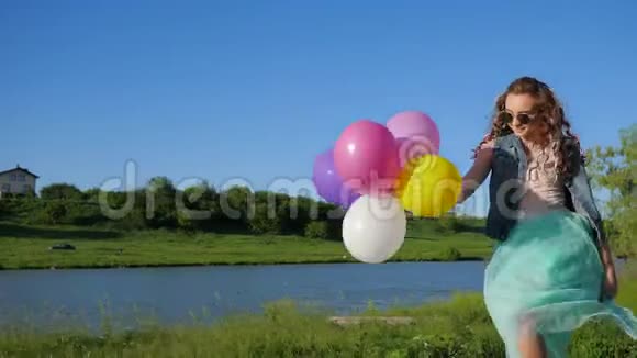 幸运的少女戴着墨镜手里拿着气球在草地上的河岸上散步视频的预览图