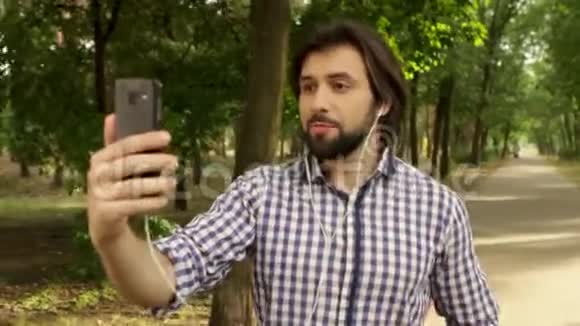有魅力的人正在公园的路上走着拿着电话对着脸他戴着耳机有人在说话视频的预览图
