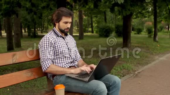 有胡子的人坐在公园的长凳上和拉普罗一起工作他正在打字喝杯咖啡人在滑视频的预览图