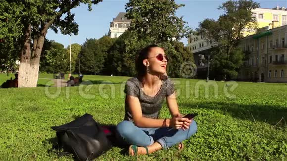 美丽微笑的年轻女孩坐在公园的草地上在电话里听她最喜欢的旋律视频的预览图