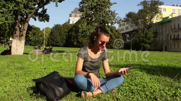 美丽的年轻女孩坐在公园的草地上在电话里听她最喜欢的曲调视频的预览图