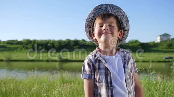 可爱的微笑男孩戴着帽子在湖附近的绿色草地上喝着玻璃水视频的预览图