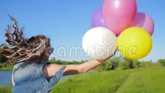 一个年轻时尚的女孩头发上长着彩色的卡尼卡龙手臂上有球跳在河边的绿色草地上视频的预览图