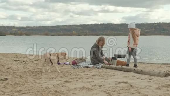 一位穿着外套的年轻女士坐在河边的海滩上大海野餐从热水瓶里倒热茶旁边是一个视频的预览图