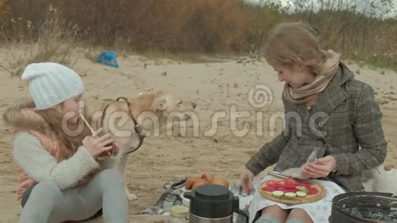 年轻的女人穿着外套一个卷发女孩妈妈和女儿坐在河边的海滩上大海野餐视频的预览图