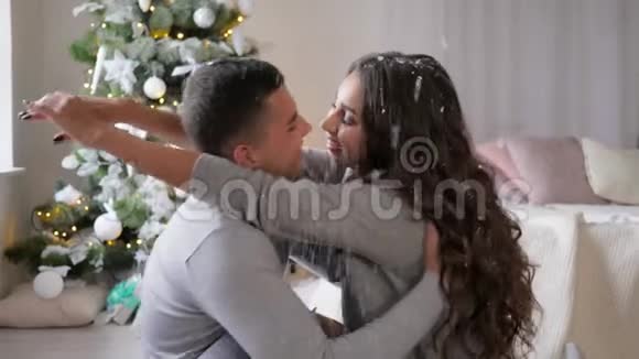 幸福的年轻夫妇坐在房间的地板上在新年树附近的五彩纸屑下缓慢地接吻视频的预览图
