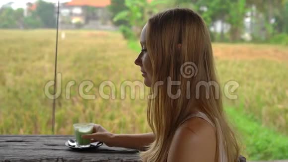 一位年轻女子在一家热带咖啡馆喝绿茶抹茶拿铁时的慢镜头观看稻田视频的预览图