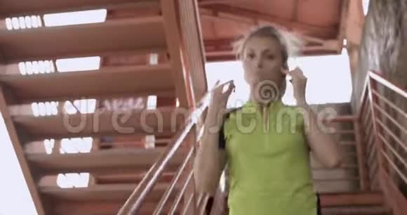 年轻的金发女郎穿着健身服走下楼梯为跑步训练热身夏日阳光明媚工业绿色视频的预览图