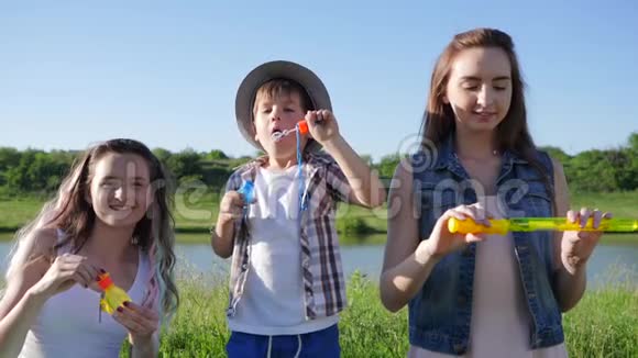 孩子们玩得很开心情绪激动的姐妹和戴帽子的小男孩在湖边的草地上对着蓝天吹肥皂泡视频的预览图