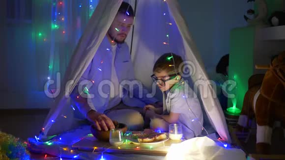 晚上吃饭爸爸和男孩一起吃面包店的产品坐在装饰有花环的帐篷里在儿童房视频的预览图